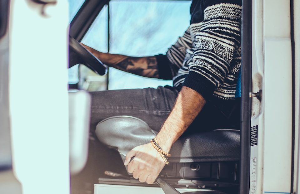 Man with tattoo in a patterned jumper sitting in a vehicle, holding the door handle, partial driver's view