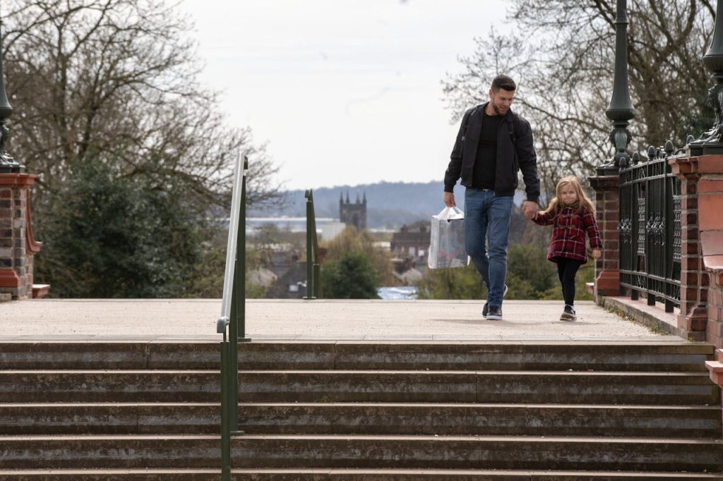 Father and daughter walking by steps in park holding hands, man carrying shopping bag, trees and town view in the background